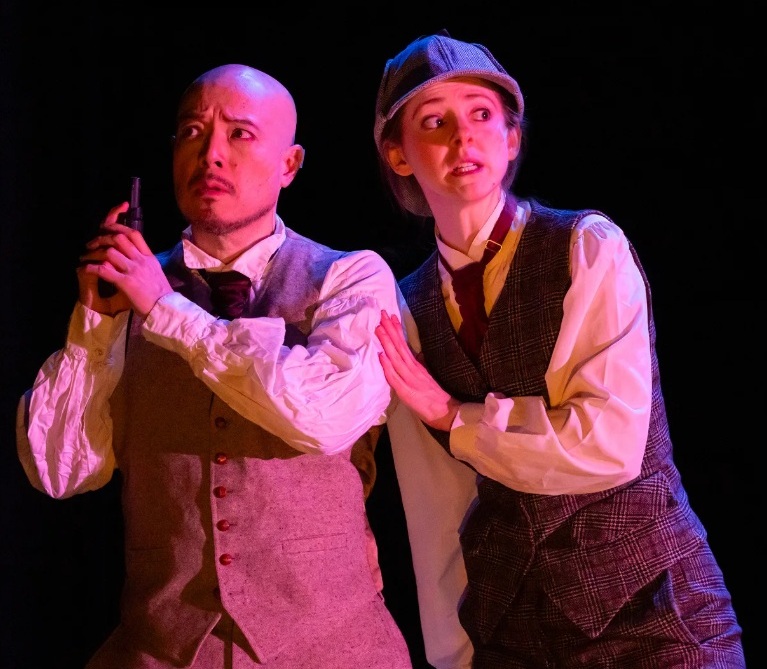 A bald man in a tie and beige vest holding a gun pointing upward, and a woman in a deerstalker wearing a tie and a dark gray vest.