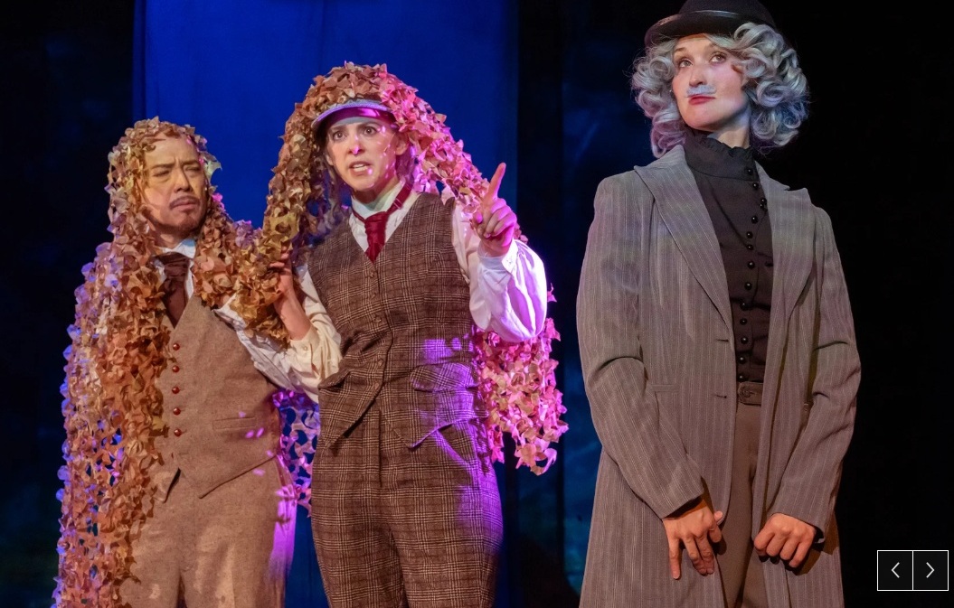 A man in a tie and vest wearig a long curly wig, a woman in a brown plaid vest also wering a long curly wig and a woman wearing a long jacket disguised in a moustache.