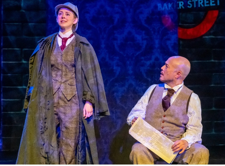 A woman in a deerstalker and long gray coat standing next to a bald man in a tie and beige vest sitting down holding a newspaper.
