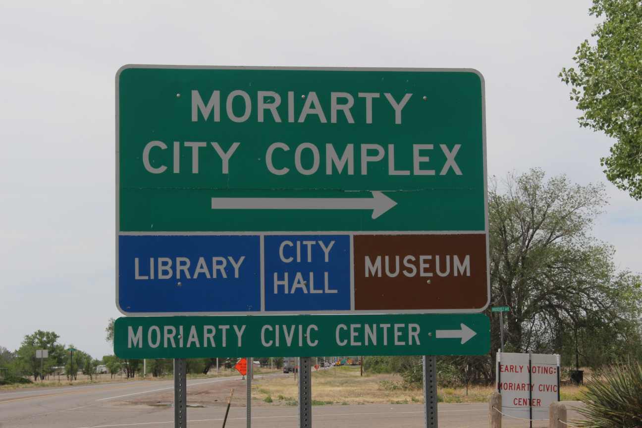Highway sign that says Moriarty City Complex with an arrow pointing right, then below that says Library, City Hall and Museum, and below that Moriarty Civic Center with an arrow to the right.